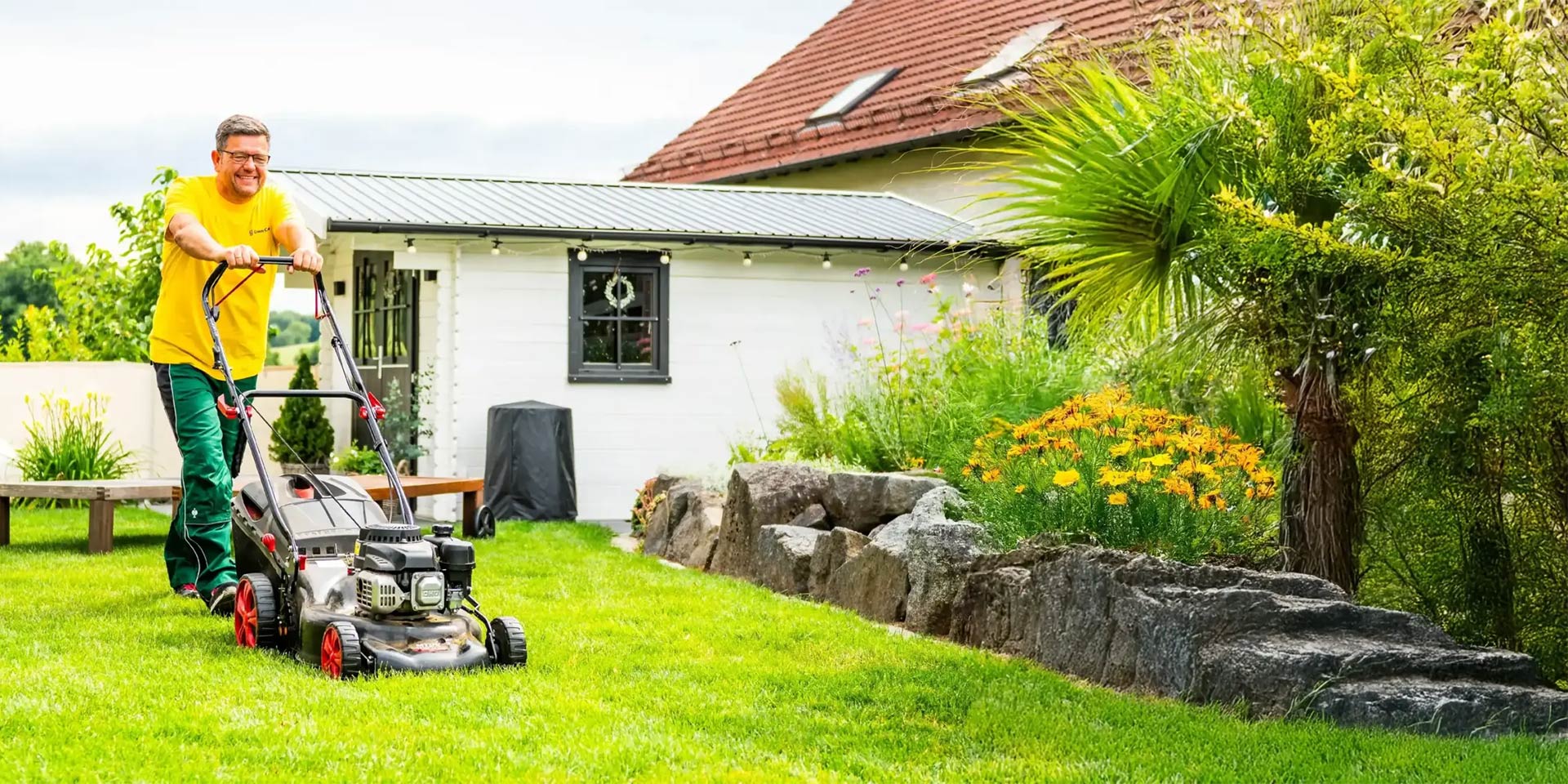 Mann in gelbem Shirt mäht mit einem Rasenmäher den Rasen im Garten vor einem Haus
