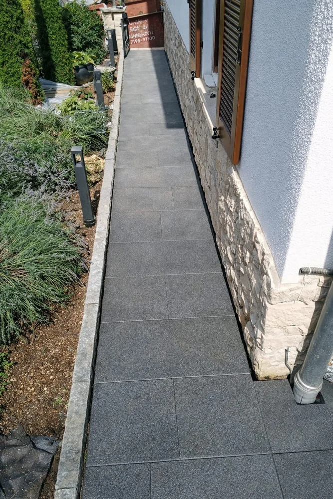 Plattenweg neben derselben Hausfassade mit bepflanztem Beet und niedrigen Leuchten